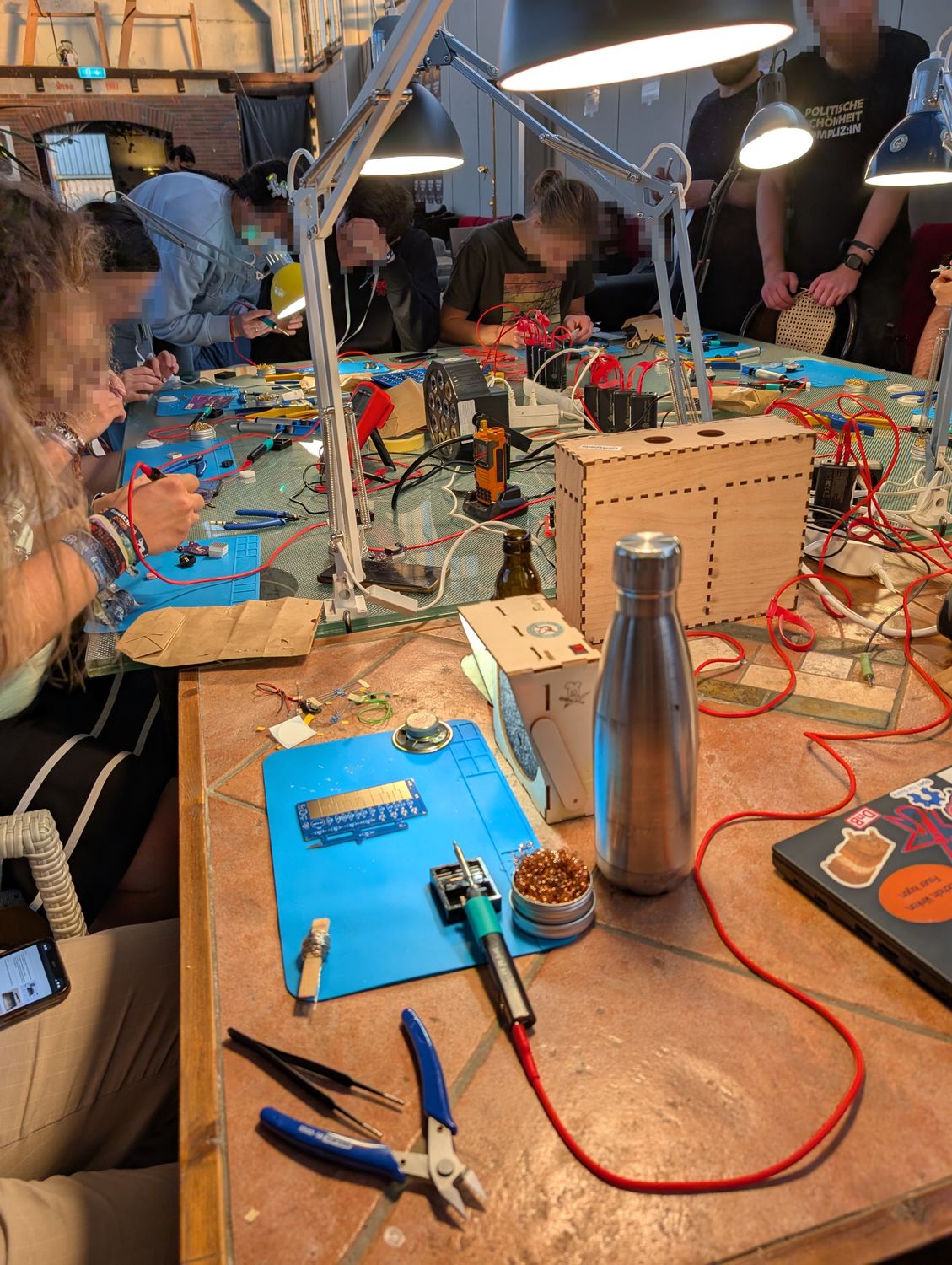 Ein großer Tisch ist als Lötarbeitsplatz aufgebaut. Daran sitzen viele Menschen und löten.